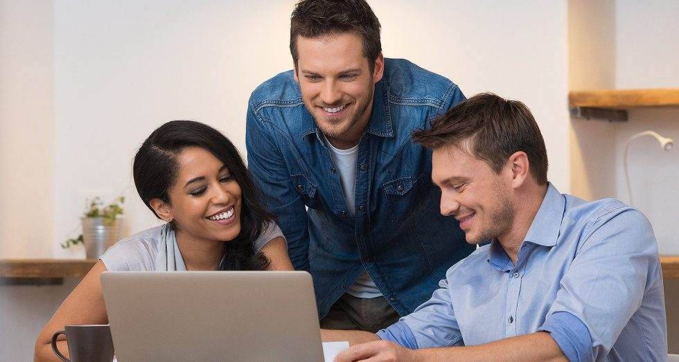 Three people, two men and a woman look at a laptop smiling in what seems like an information sharing moment. On of the men in denim jacket is standing.